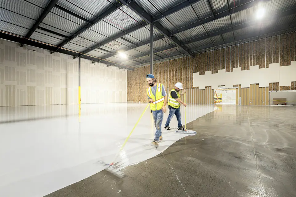 Two epoxy floor installers spreading a gray coat with squeegees over prepared concrete (stock photo)